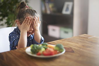 Little girl hiding from a plate of vegetables