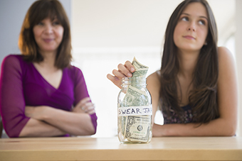 Teen girl putting money into a jar labeled "swear jar" with mom watching